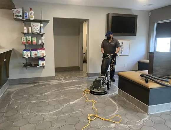 Cleaning professional using a floor machine in a commercial space with cleaning supplies on a shelf.